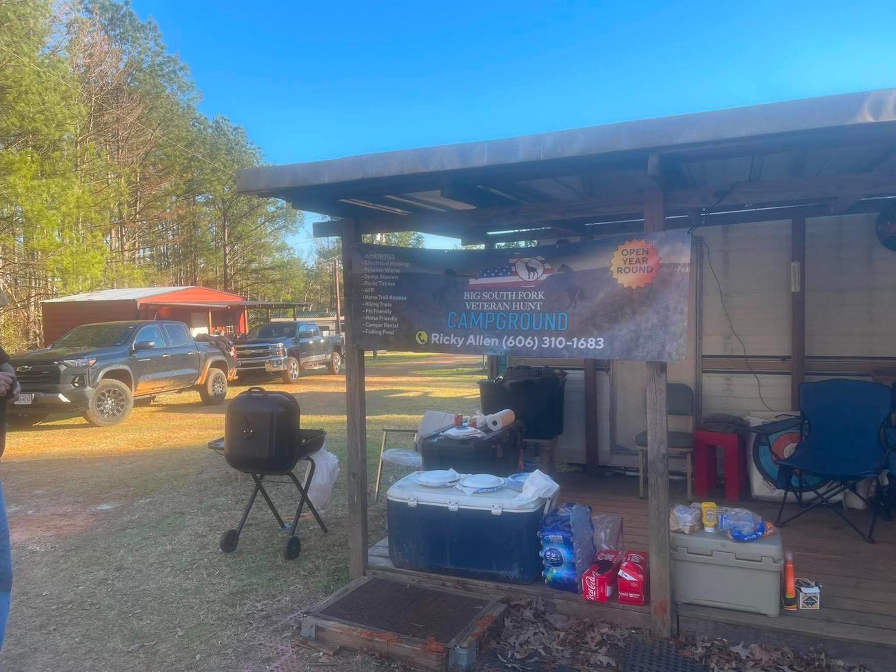 The Big South Fork Veteran Hunt Campground amenities banner on the covered deck at the campsite, with trucks parked and a grill set up.