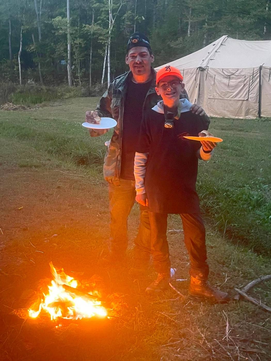 Two veterans sharing a plate of food by the campfire at dusk.